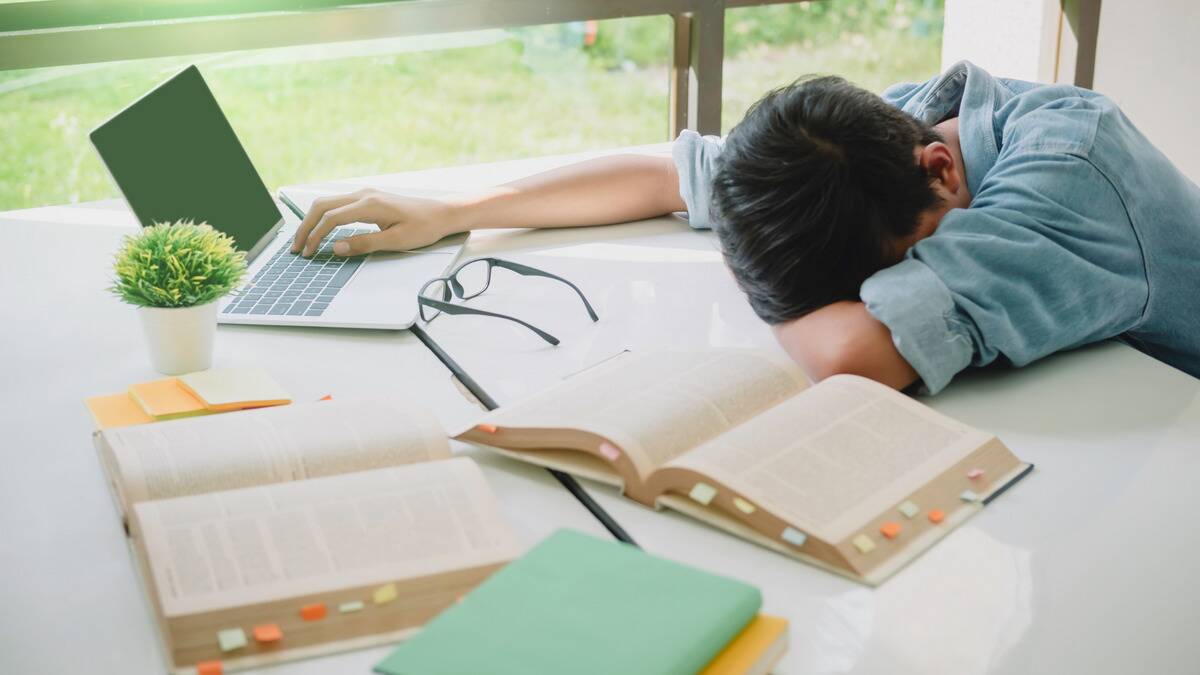 A student face down on his desk, head in his arm, open books and a laptio scattered on the desk surrounding him. 