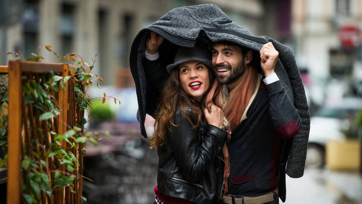 A man and woman standing outside in the rain, the man lifting his jacket up to cover them both.