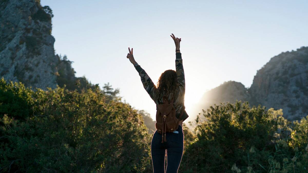 A woman facing away from the camera, arms up with her hands throwing peace signs as she stands in the sun.