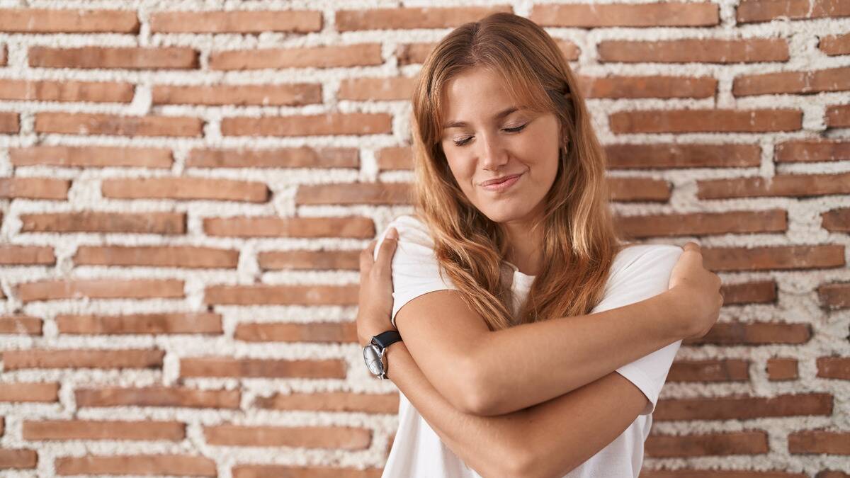 A woman standing in front of a brick wall, her eyes closed and smiling as she hugs herself.