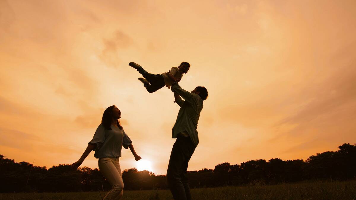 A father lifting his young son into the air, the mother nearby with them, all smiling outside as the sun sets.