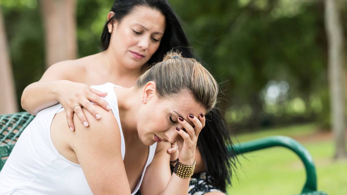 A woman sitting on a bench outside, hunched over, her head in her hand. Her friend is sitting next to her with a comforting hand on her shoulder.