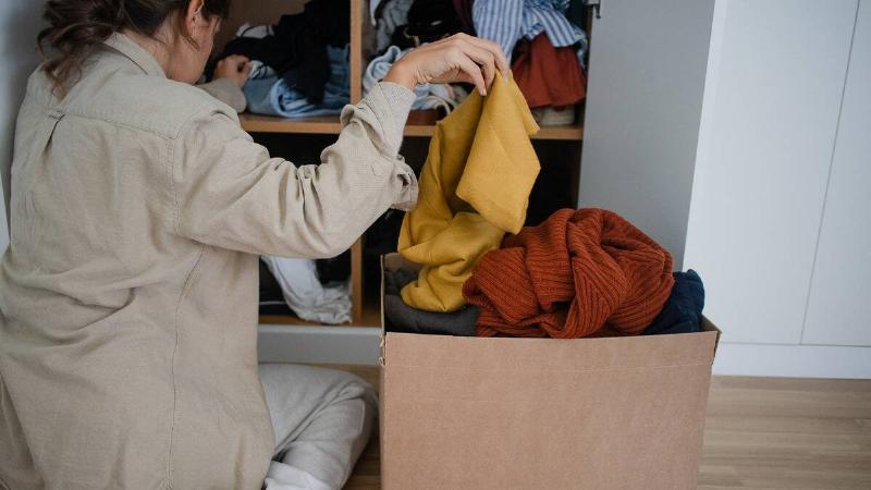 A woman is sitting by her closet, sorting through her clothes and putting some in a box.