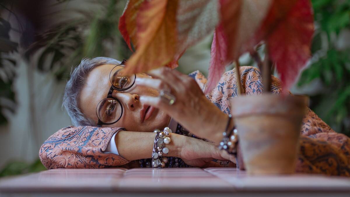 A woman laying her head on her arm against a table, reaching out to touch the leaves of a houseplant, looking stressed and forlorn.