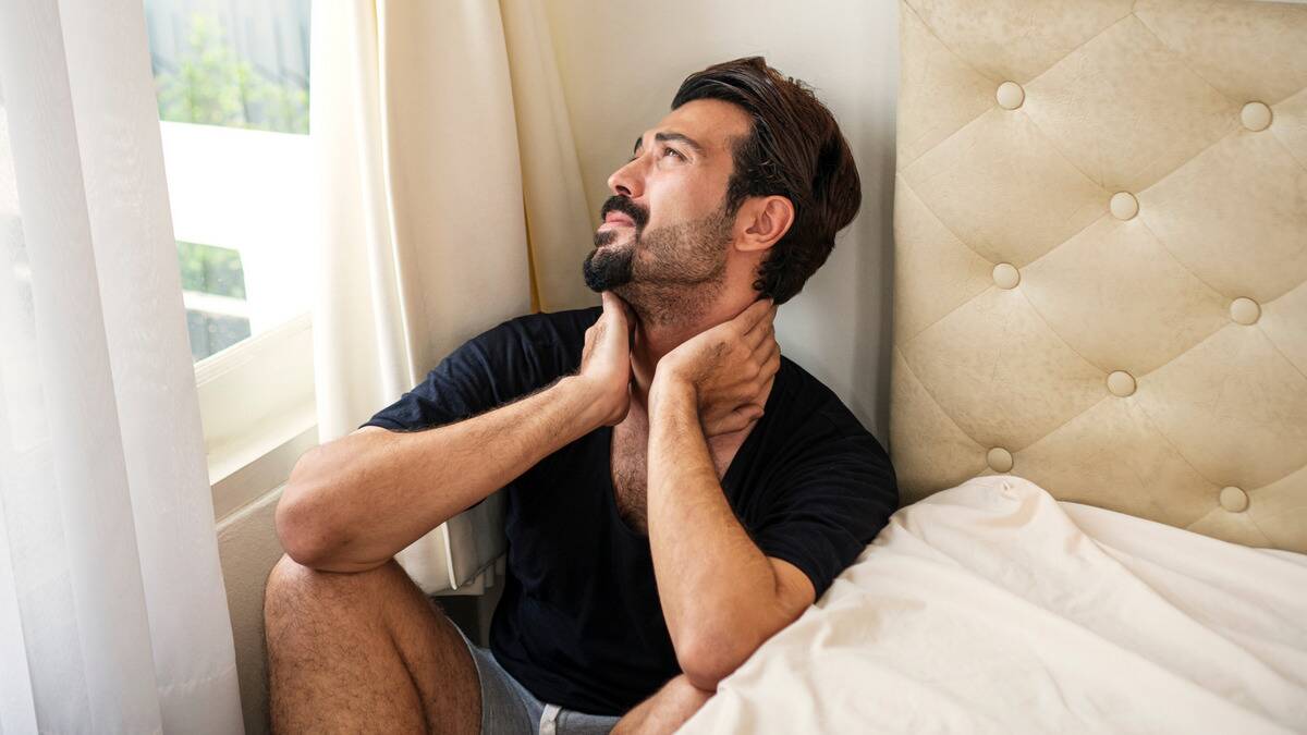 A man sitting between the side of his bed and the wall, hands on his neck, looking out the window with a stressed expression.