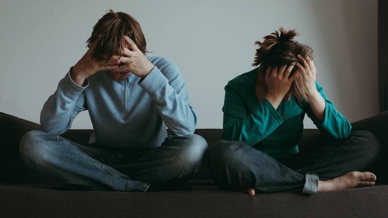 A couple sitting side by side on their bed, both cross-legged, both with their head in their hands.