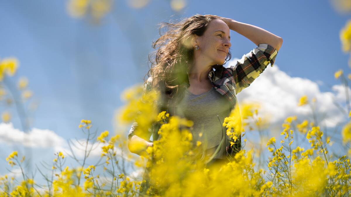 A low-angle shot of a woman standing amid some yellow flowers, her hand in her hair that's being blown back by the wind.