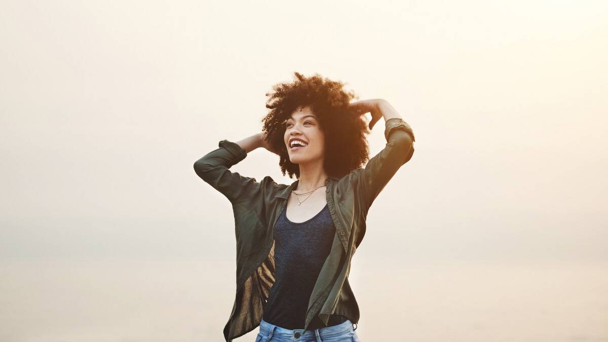 A woman standing in the hazy sunshine, smiling with her hands up by her hair.