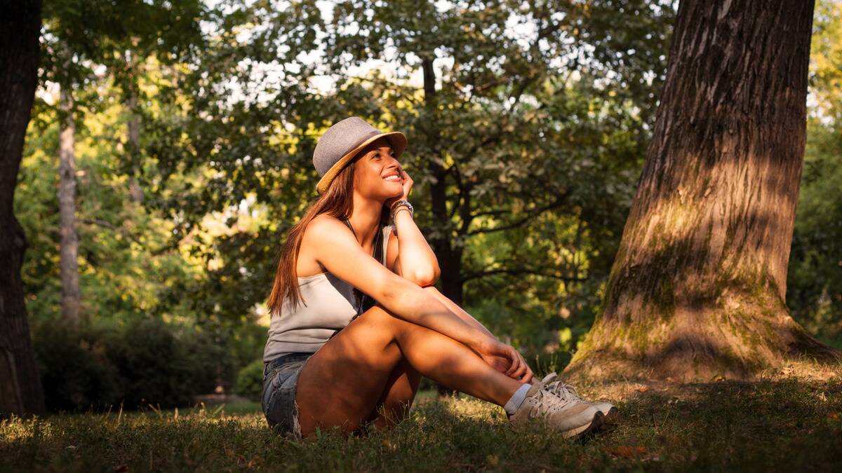 A woman sitting beneath a tree, her knees drawn up, her shin in her hand, smiling as she looks up at the sun shining down on her.