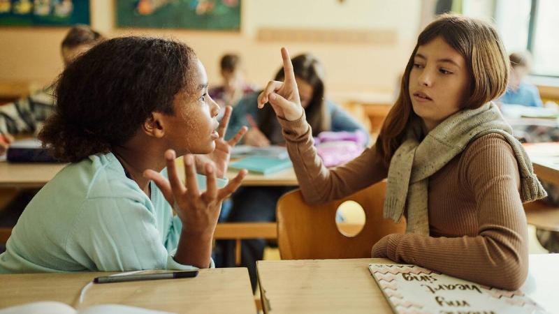 Two young girls in class, one looking frustrated as the other raises a finger to interrupt her.