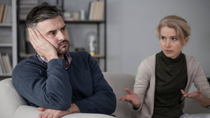 A man looking tired, leaning his cheek in his hand, while his wife sits next to him complaining.