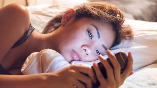 A woman laying on her side in bed, looking sad or tired as she stares at her phone, the screen illuminating her face.