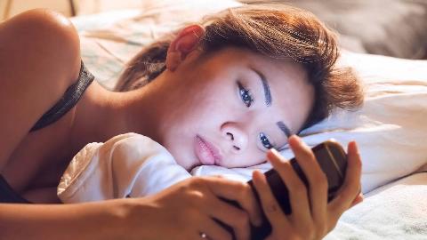 A woman laying on her side in bed, looking sad or tired as she stares at her phone, the screen illuminating her face.