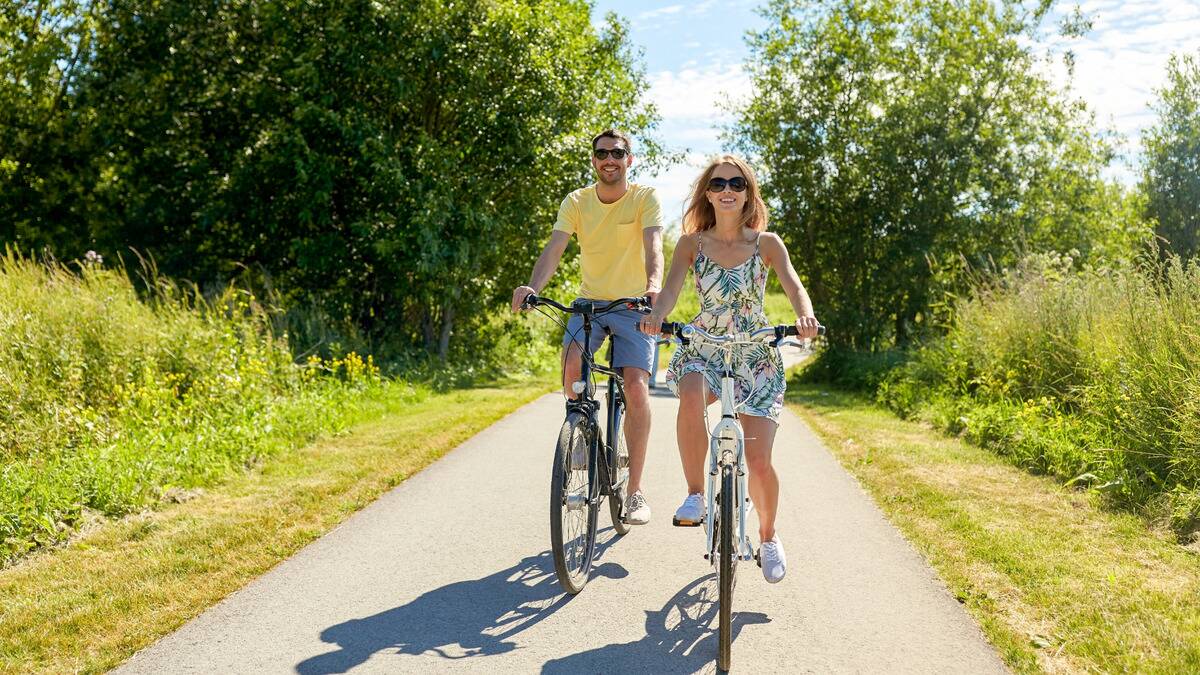 A couple riding their bicycles down a paved path on a sunny day, both smiling and wearing sunglasses.