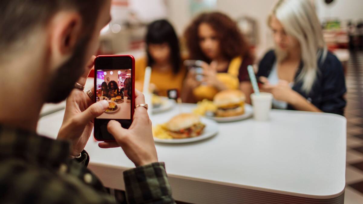 A man taking a photo of the restaurant table he's eating at, including the food on it, as well as the three women sitting opposite him.