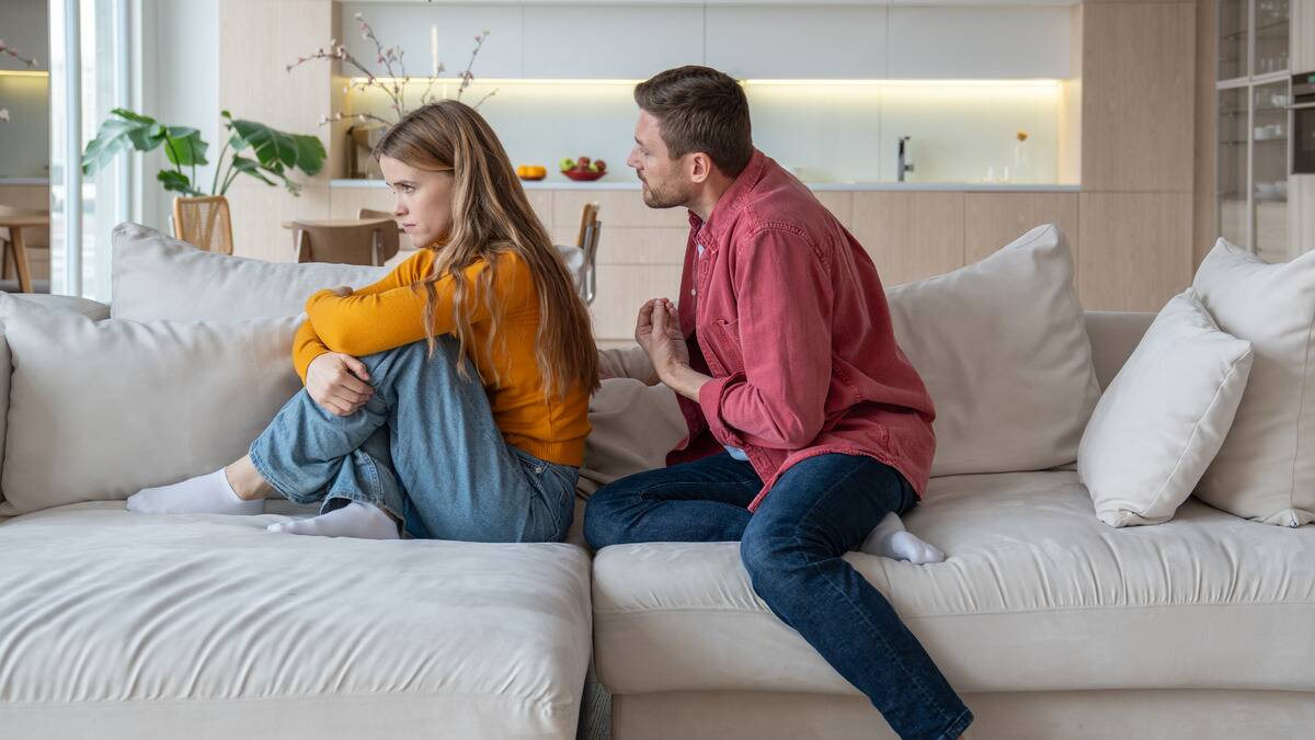 A couple on their couch, the woman facing away from the man, her knees drawn up to her chest, looking annoyed. The man is behind her pleading, trying to get her to talk to him.