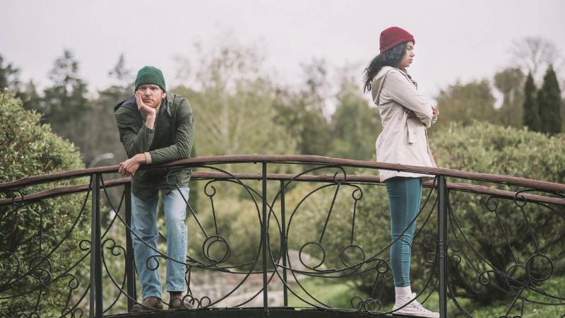 A couple standing apart on a small bridge in a garden, the woman facing away from the man with her arms crossed, the man leaning against the railing, chin in his hand.