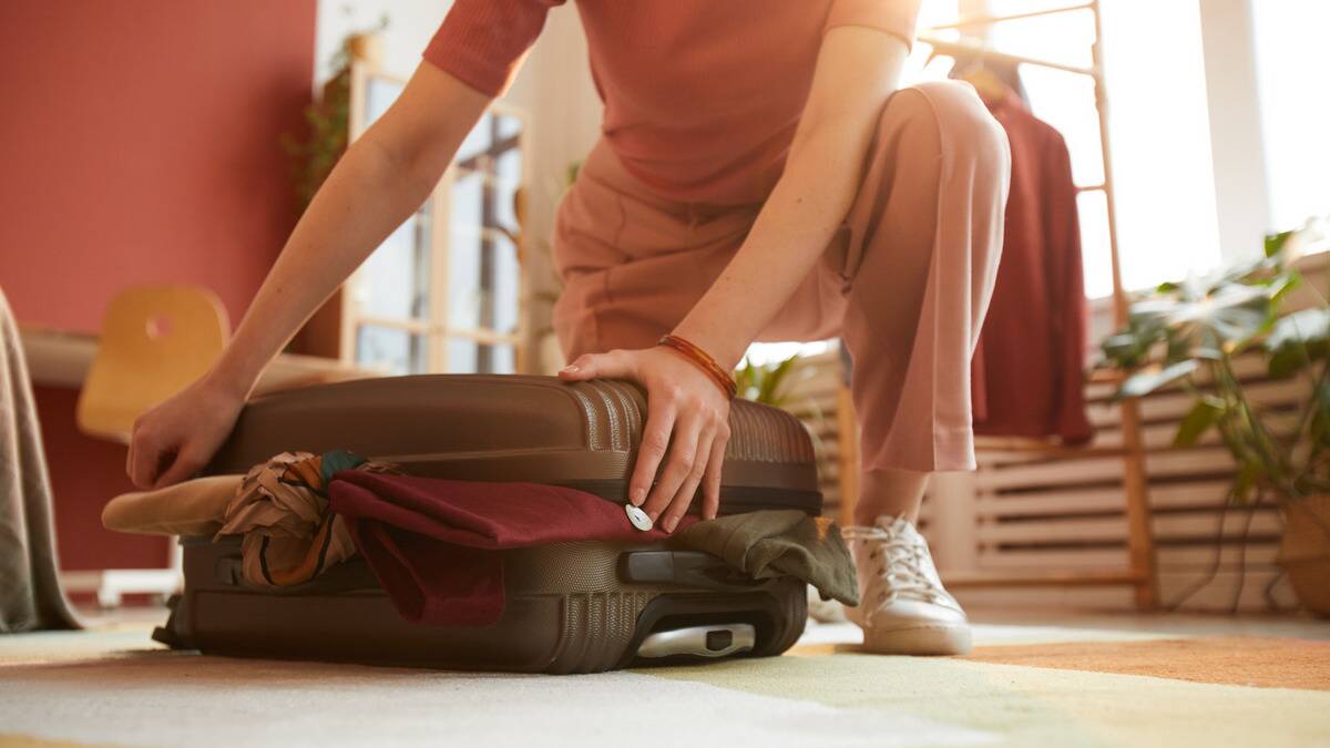 A close shot of a woman kneeling on the ground trying to push down a small overpacked suitcase.