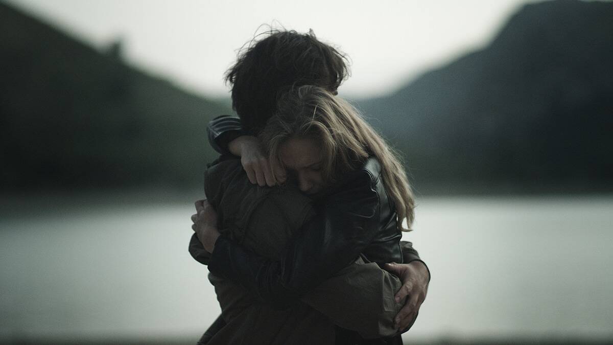 A dark photo of a couple hugging tightly, the woman, whose face we can see, looking upset or distressed.