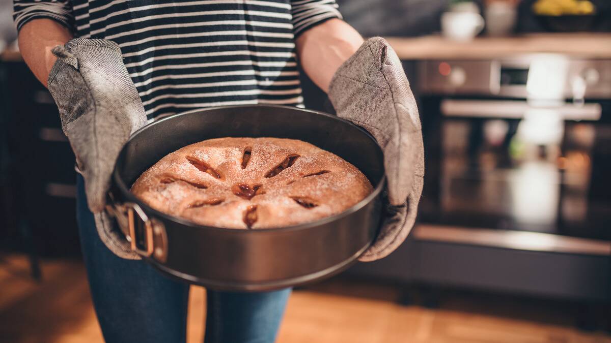 Someone in large grey oven mitts carrying a freshly baked pie in a springform pan.