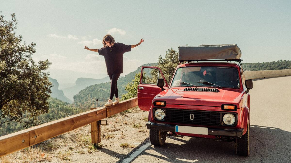 A woman walking along a railing next to a road at a cliffside, her car parked next to her with the passenger door open.