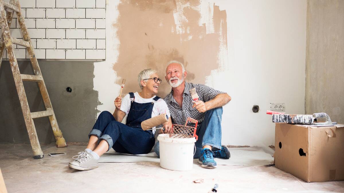 A couple sitting against a wall they've started painting together, both smiling as they take a break from working.