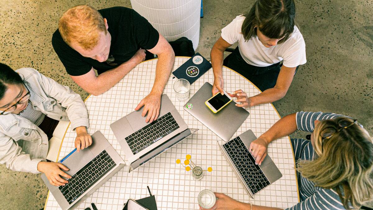 An arial shot of four friends working around a circular table, three out of the four of them typing on open laptops, two turned to look at each other.