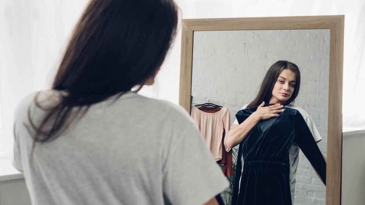 A woman looking at herself in a large mirror as she holds up a dress to her body.