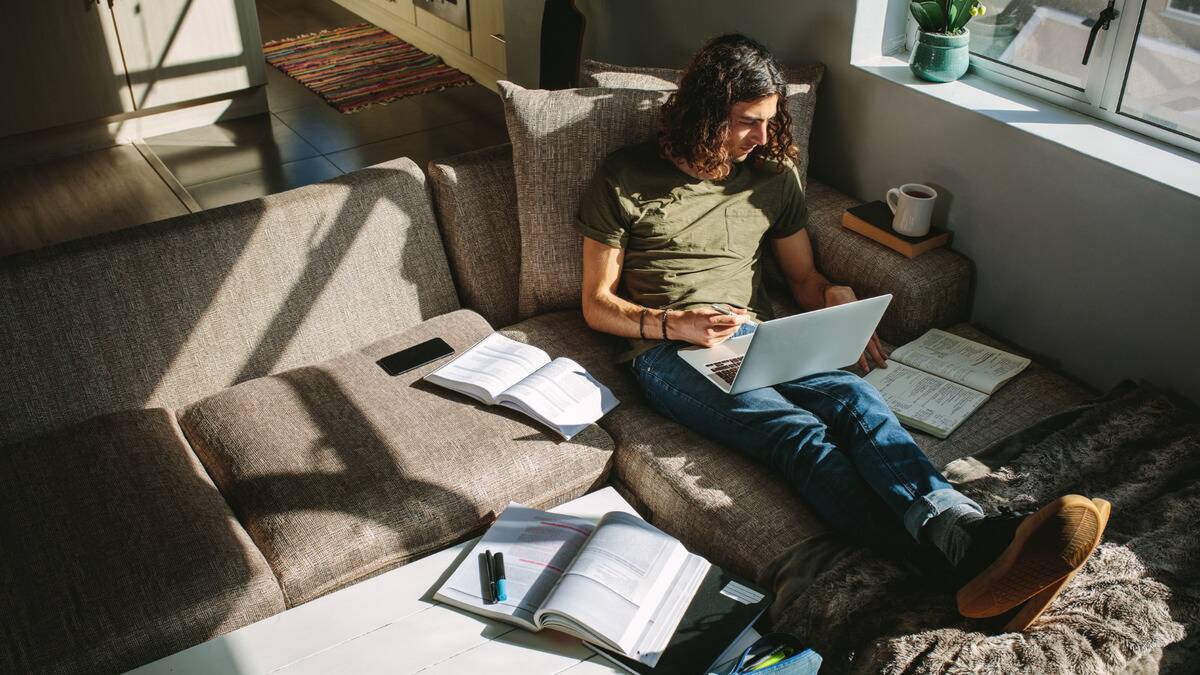 A man sitting on his couch, legs kicked up, laptop in his lap while he's surrounded by textbooks.