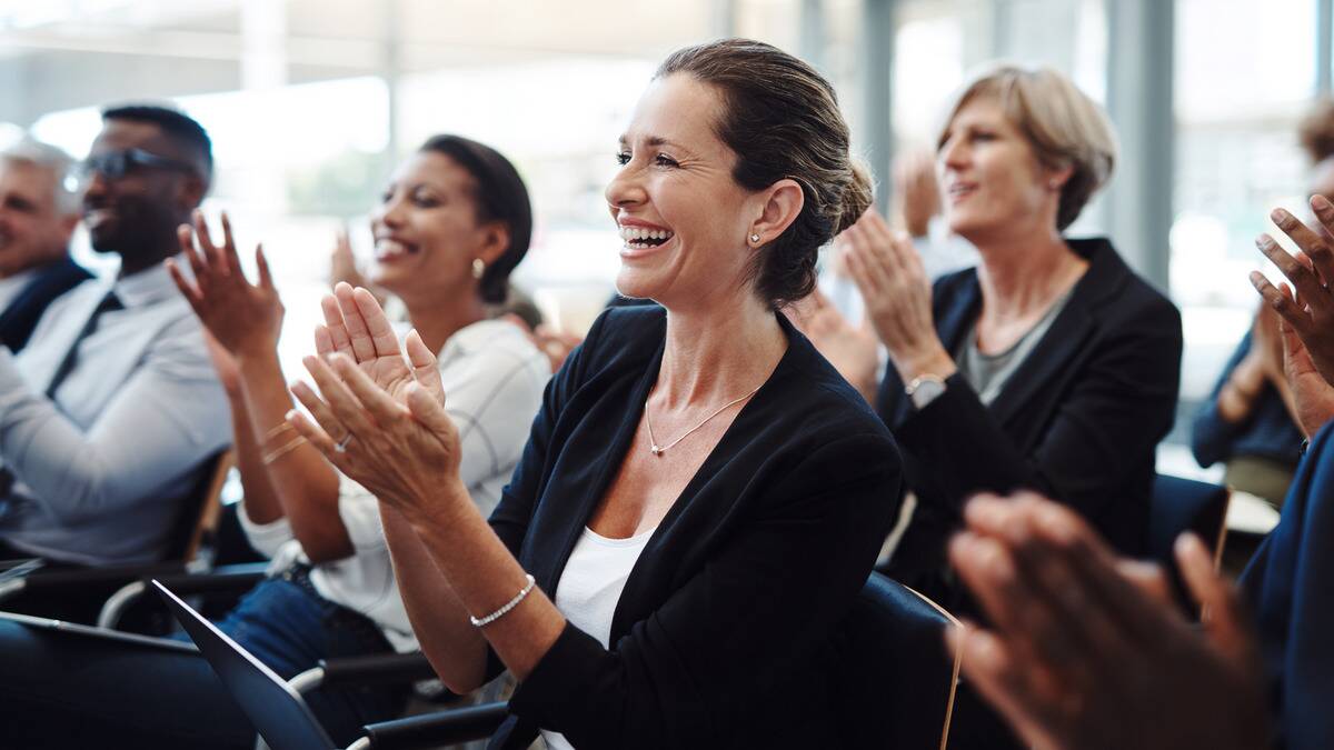 A woman sitting in the crowd of a work meeting, smiling and applauding along with her coworkers.