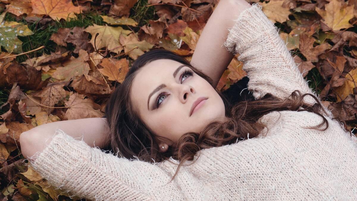 A woman laying on the ground in a pile of autumn leaves, hands behind her head, looking upward with a concerned, thoughtful expression.