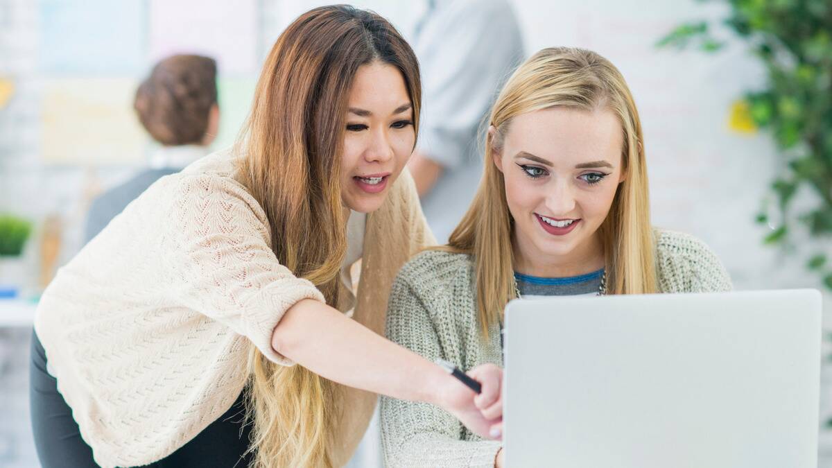 Two female coworkers, one on her laptop, the other leaning over her shoulder and pointing something out on the monitor.