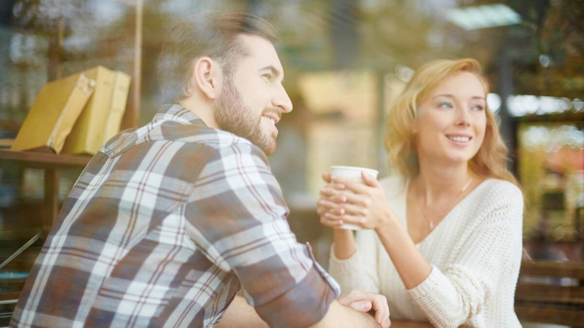 A couple sitting across from each other at a cafe, both turned to look out the window, the woman holding a coffee cup with both hands.