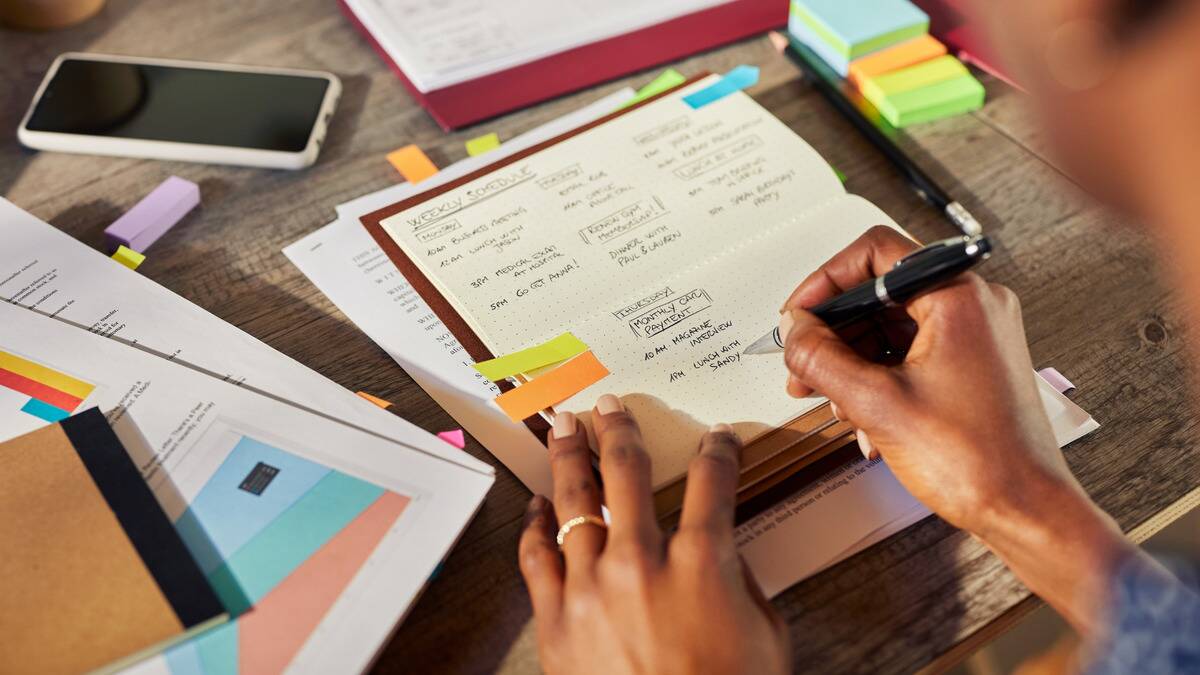 An over-the-shoulder shot of someone writing out their week's schedule in a planner, surrounded by notes and colorful post-its.