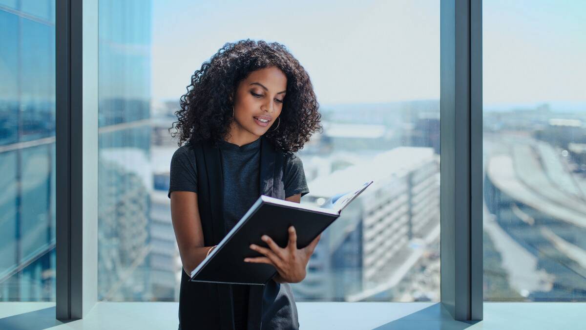 A woman at work, standing in front of a large office building window, writing in a large folder she has open in front of her.