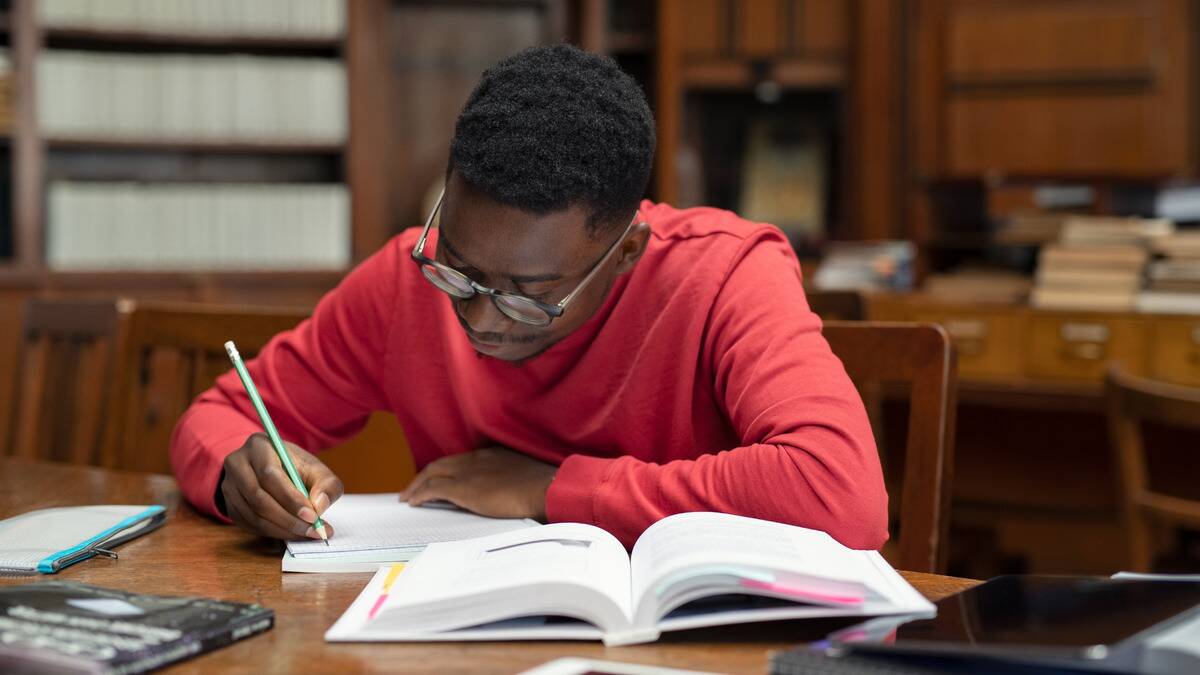 A man sitting at a library desk, hunched over a notebook he's writing in, a text book open in front of him.