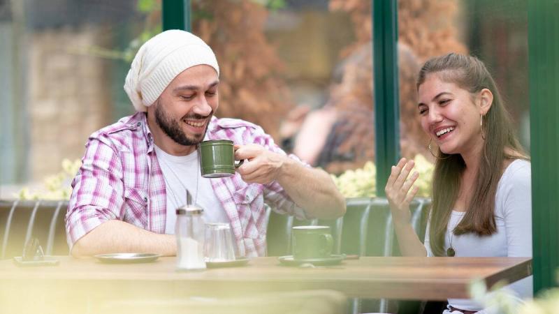 A man and woman hanging out at a cafe, both smiling as they chat.