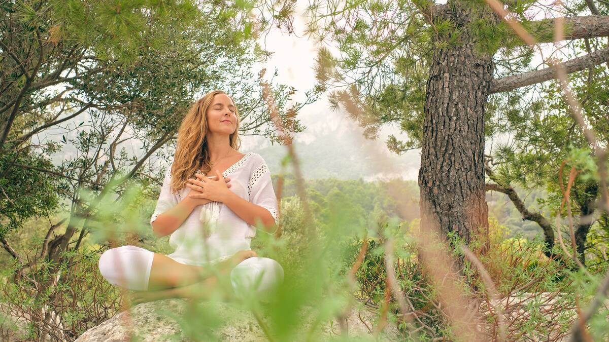 A woman sitting outside on a rock, legs crossed in a yoga pose, hands over her heart, eyes closed, smiling peacefully, shot through blades of grass.