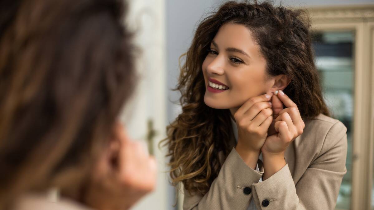 A woman smiling as she puts an earring while looking in the mirror.