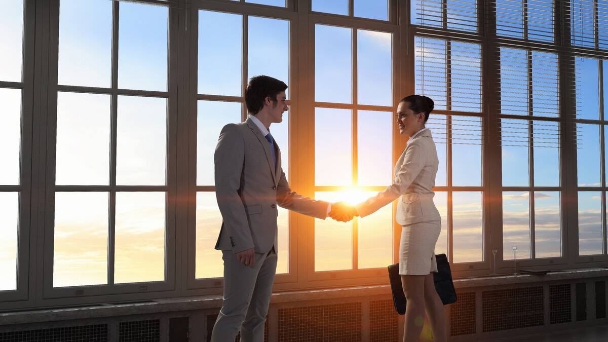 Two business professionals shake hands as the sunrise shines directly behind them.