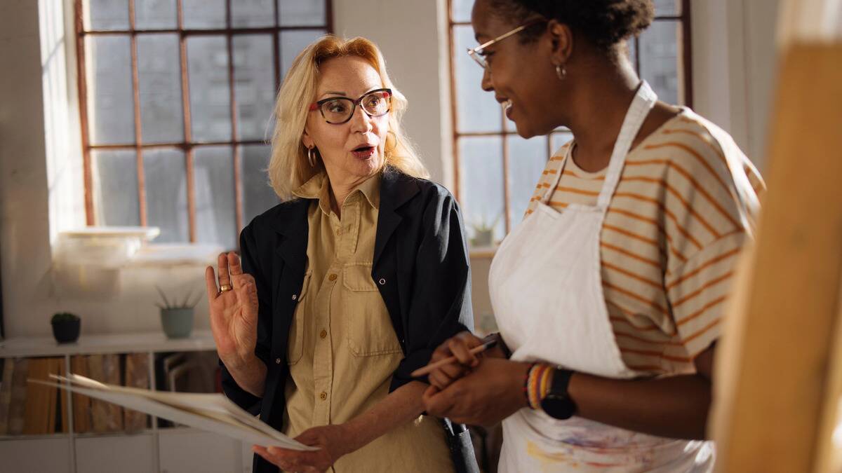 A woman giving artistic critique to a younger artist, holding her large sketch places, the artist smiling as she listens.