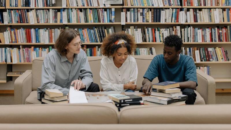 Three friends sitting on a couch side by side in a library, the table in front of them stacked with books, all examining something in particular.