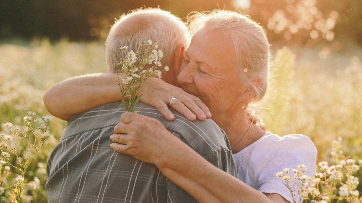 A senior couple standing outside in the sunshine and hugging, the woman holding a small bundle of delicate wildflowers.