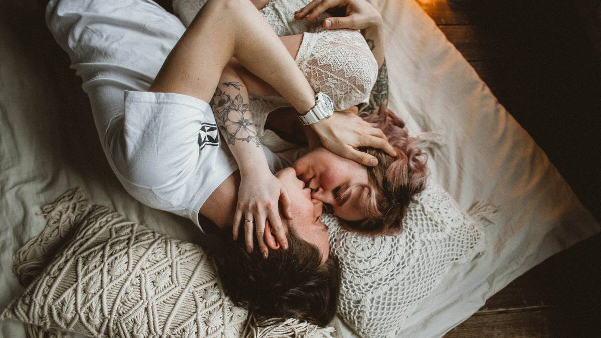 An upside down photo of a couple laying together in bed, hands on each others cheeks, kissing gently.