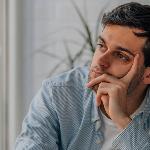 A man sitting indoors, a hand on his cheek, looking dejected.