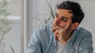 A man sitting indoors, a hand on his cheek, looking dejected.