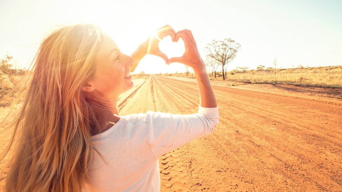 A woman standing outside in the sunlight, making a heart with her hands, looking off to the side as she smiles.