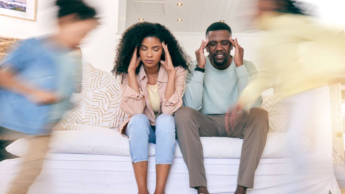 A couple sitting side by side on a couch, hands to their temples, looking stressed as children run around them.