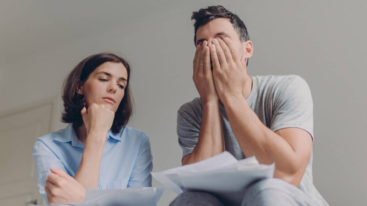 A low-angle shot of a couple sitting next to one another, both looking stressed. The woman has her chin on her hand, the man is covering his face, and both have papers in their laps.