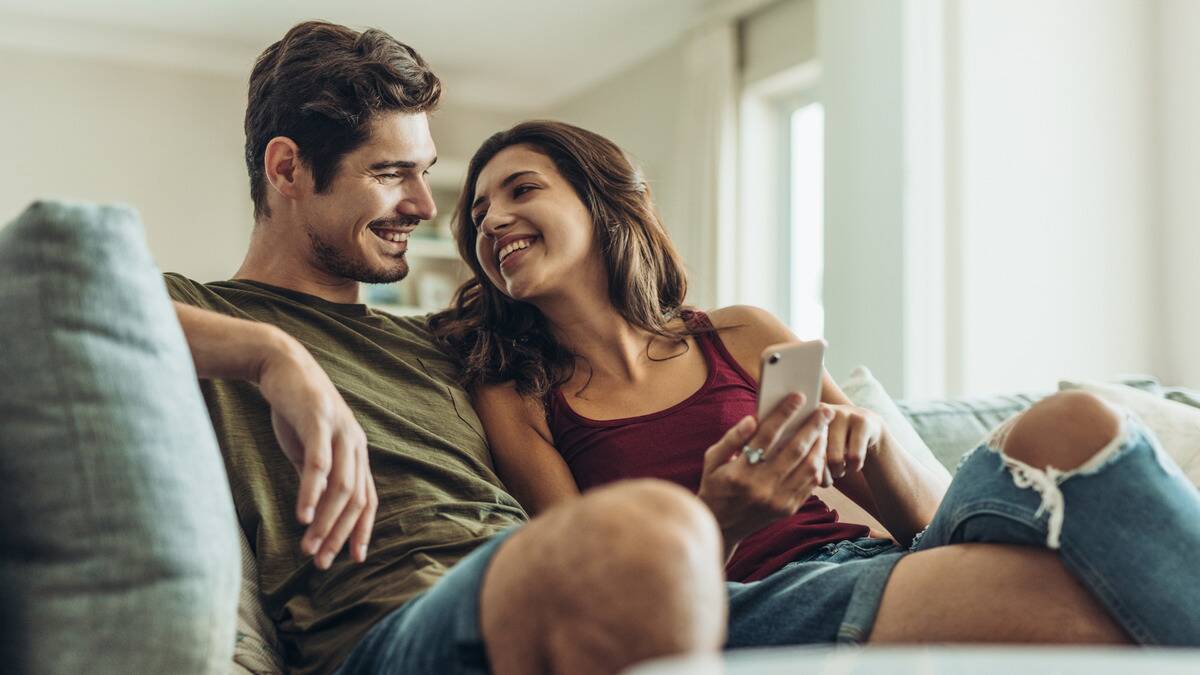 A couple sitting side by side on a couch, the woman showing the man something on her phone, both smiling at one another.